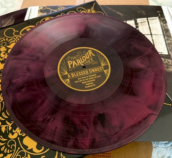 A translucent purple marbled vinyl record of A Blessed Unrest by The Parlour Trick, Meredith Yayanos’s musical project, displayed on ornate black and gold album artwork. A translucent purple marbled vinyl record of A Blessed Unrest by The Parlour Trick, Meredith Yayanos’s musical project, displayed on ornate black and gold album artwork.