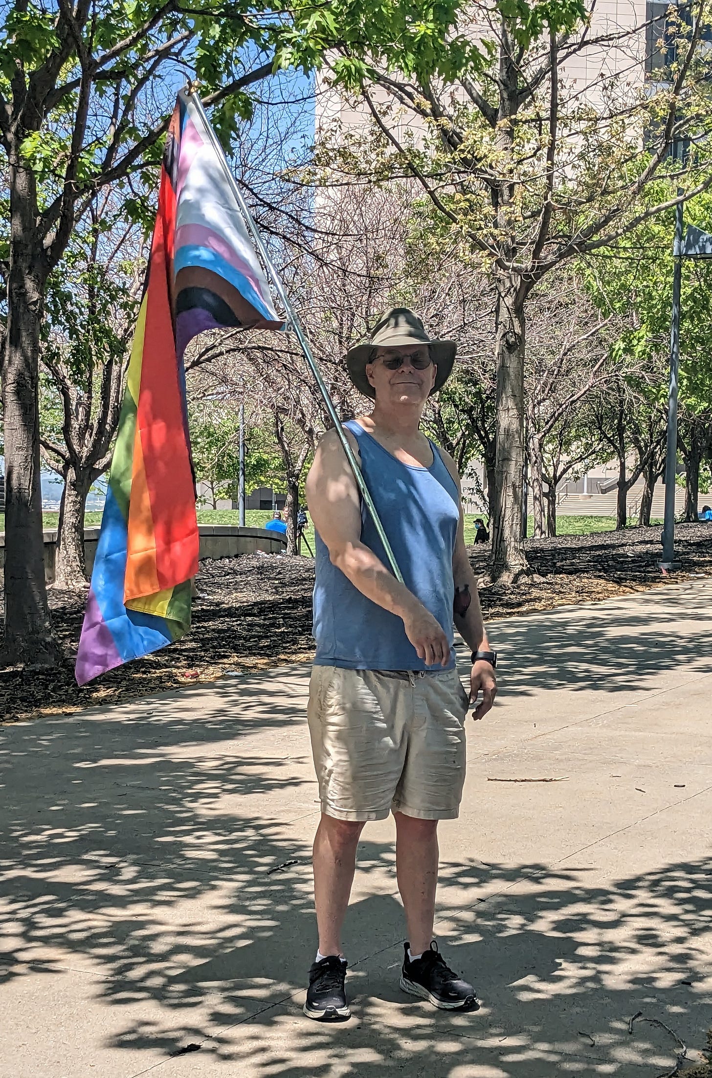 An individual in a sun hat, holding a progress pride flag and smiling proudly at the camera.