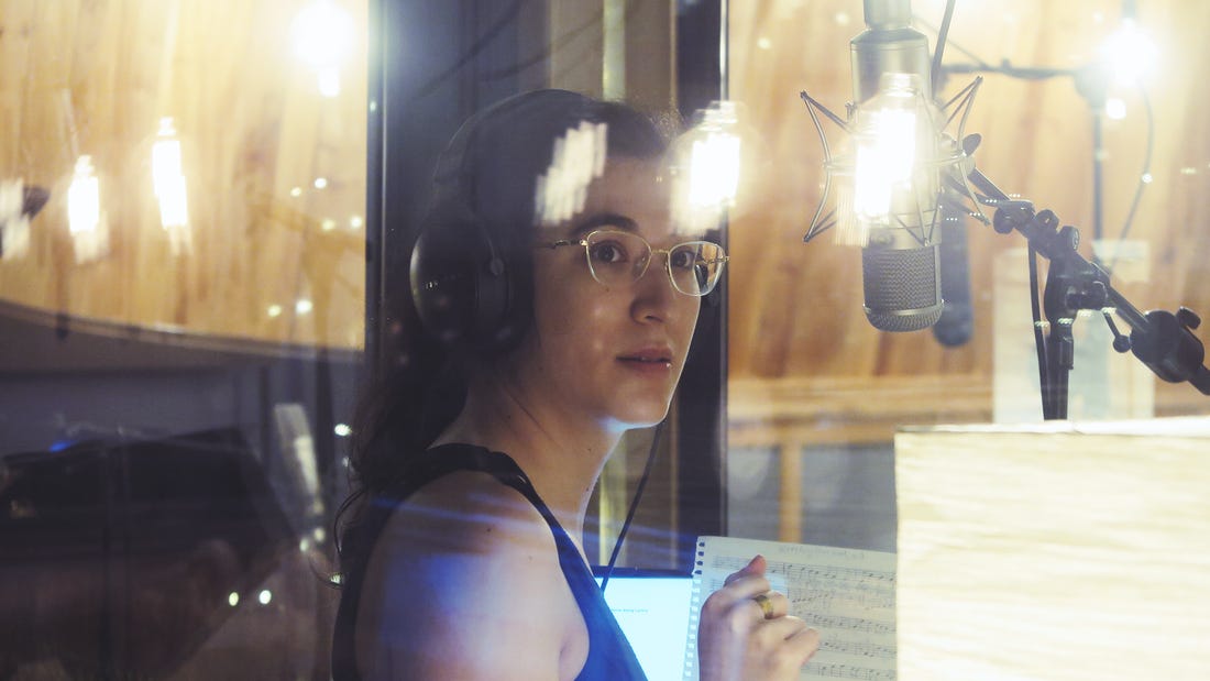Stephanie Lamprea in a studio recording booth. The lights of the studio are reflecting on the booth's glass walls.