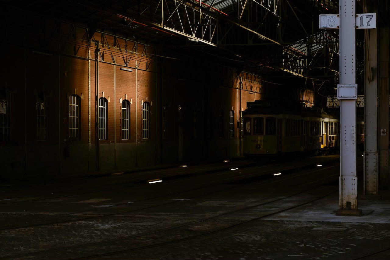 Inside a warehouse filled with deep shadow that contrasts with a few pockets of sunlight, yellow trams await.