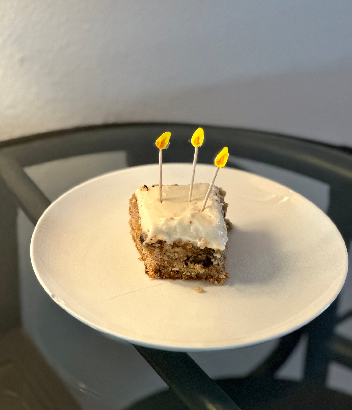 A piece of carrot cake with three toothpicks that have little pieces of paper made to look like flames on a cracked plate