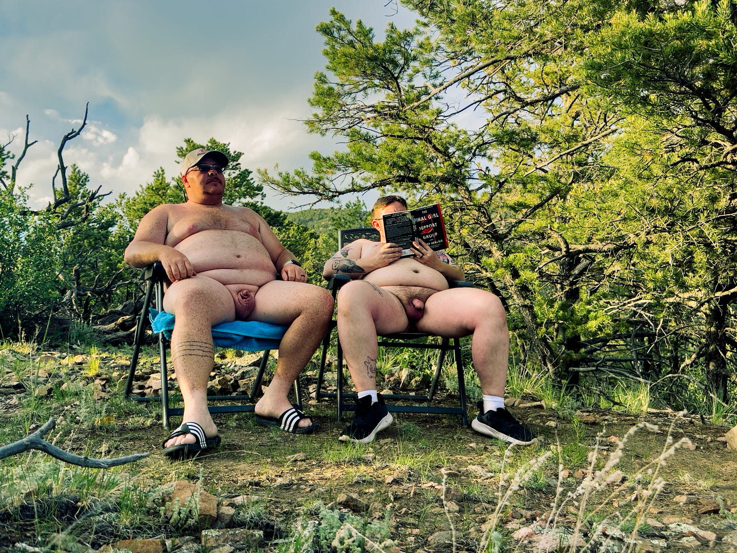 This image shows two naked men sitting in folding chairs in a rocky, wooded clearing.  The man on the left is wearing a grey baseball cap, sunglasses, and striped slide sandals while looking out toward the horizon.  The man on the right is reading a book titled The Final Girl Support Group by Grady Hendrix, wearing black sneakers and white socks.  The setting is a natural outdoor environment with pine trees and green foliage under a bright, partly cloudy sky.
