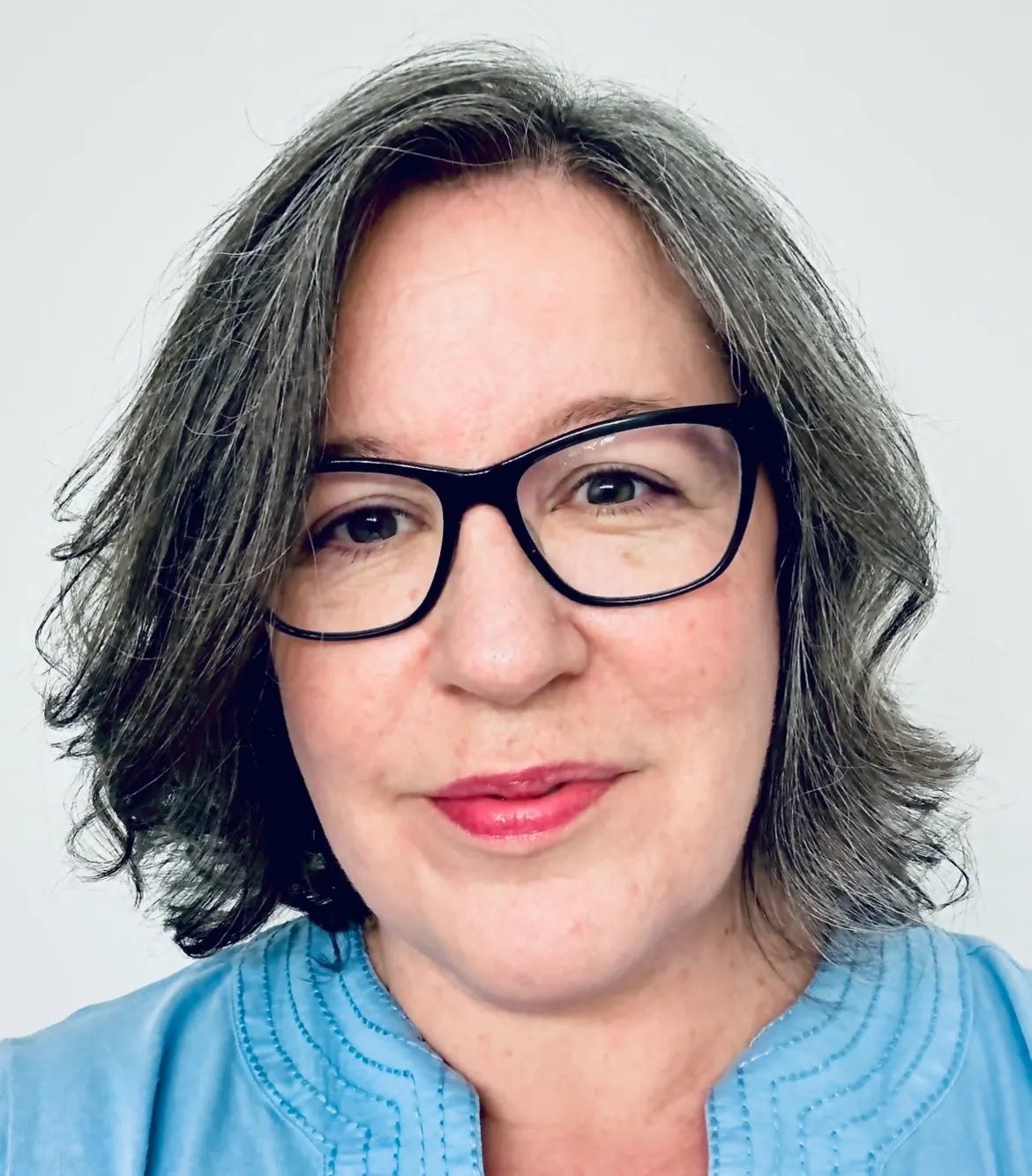 A woman faces the camera against a plain light gray background. She has shoulder-length wavy gray hair, black cat-eye glasses, pink lipstick, and a light blue blouse with stitched detailing around the neckline.