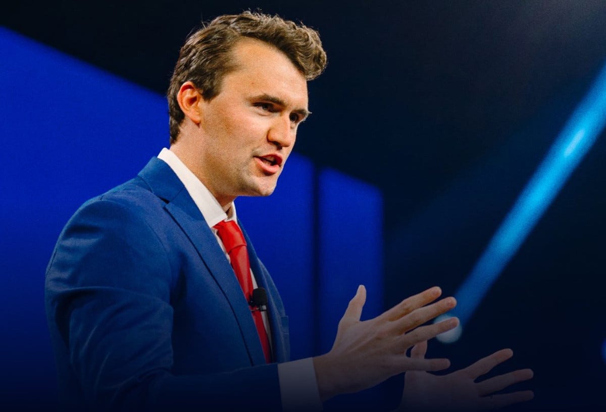Photo of Charlie Kirk facing right and wearing a blue suit, white shirt and red tie