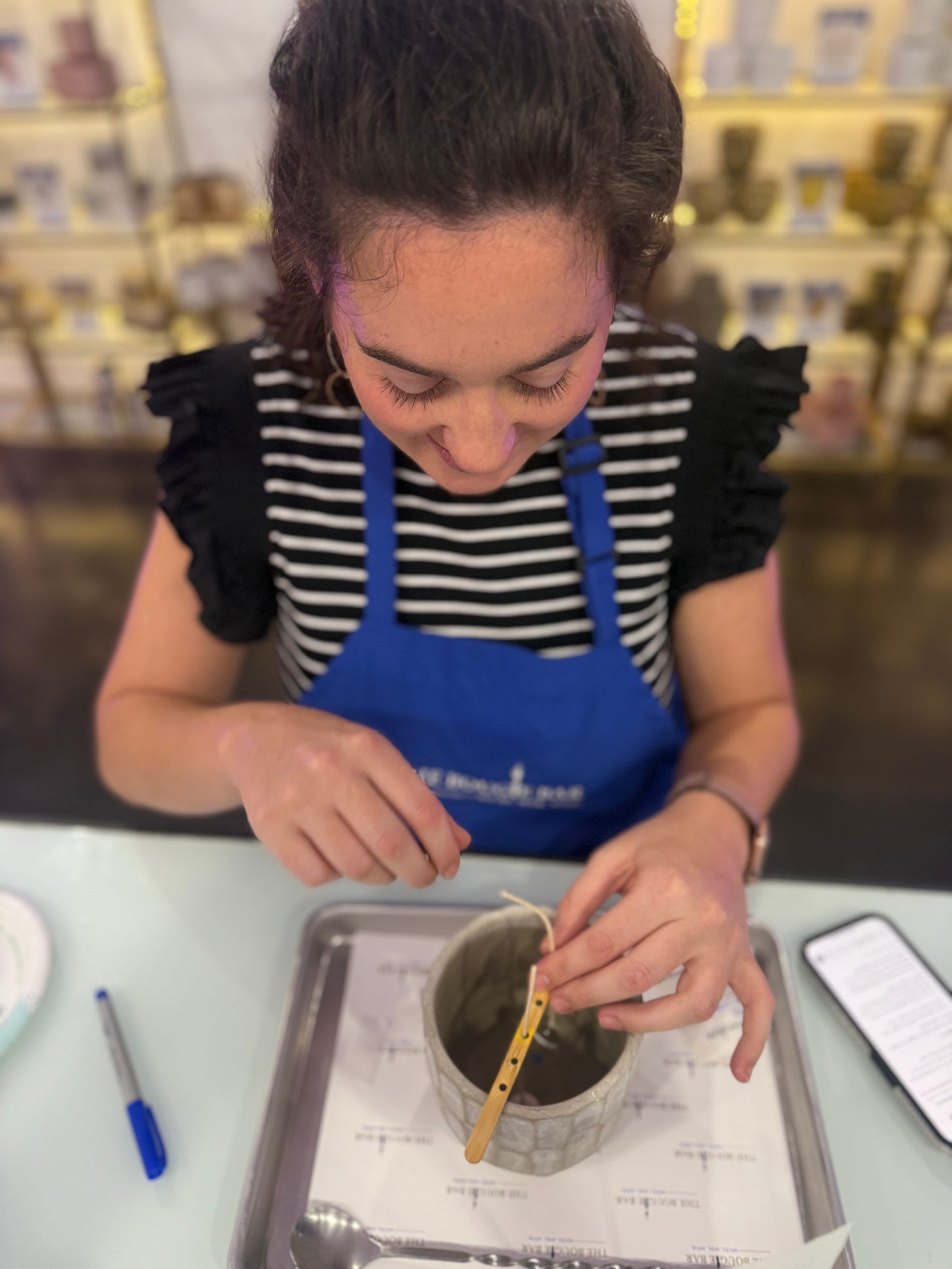 Person in striped shirt and blue Bougie Bar apron carefully measuring fragrance oil with a wooden stirrer, phone visible on table Person in striped shirt and blue Bougie Bar apron carefully measuring fragrance oil with a wooden stirrer, phone visible on table