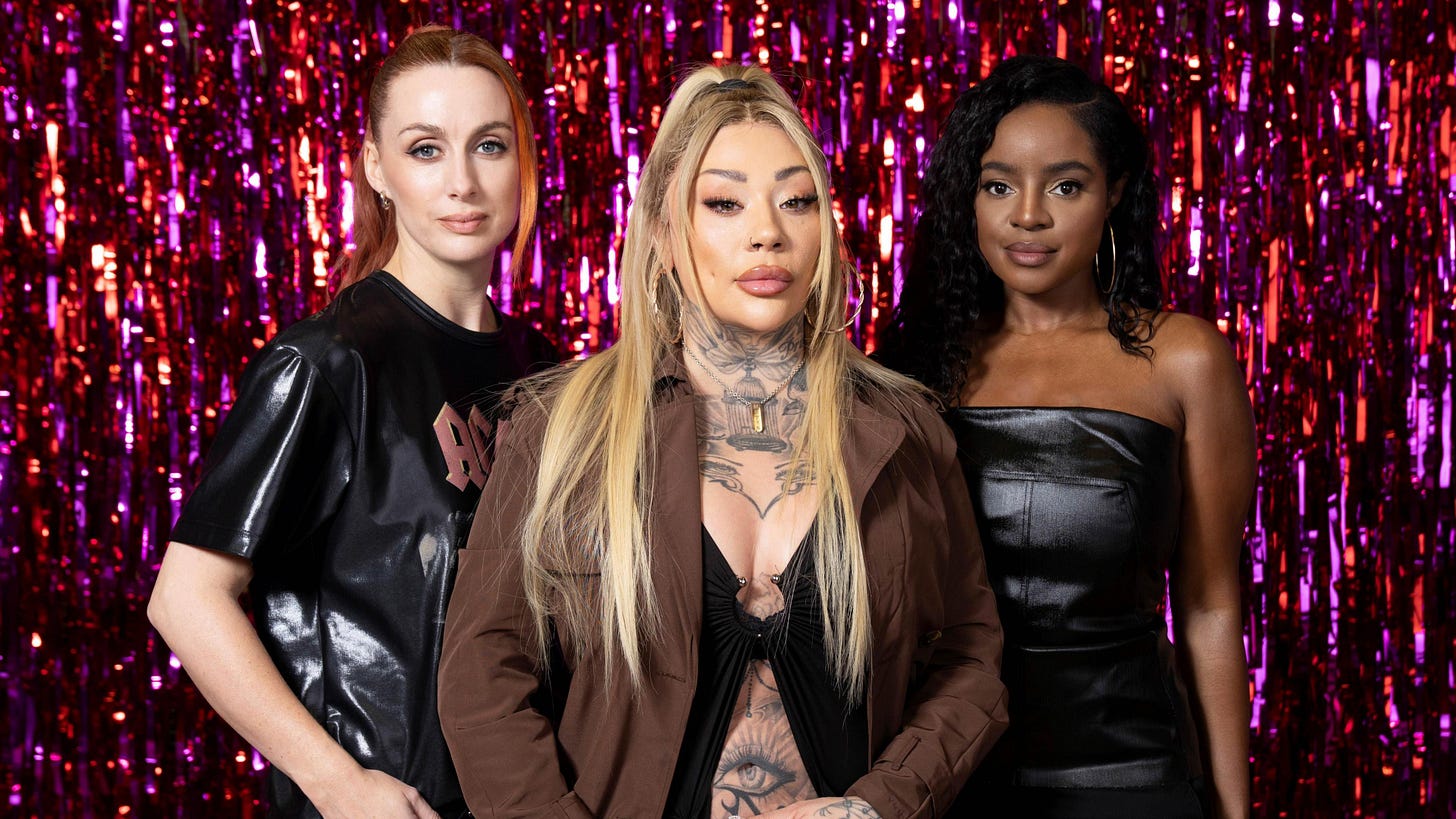 Siobhan Donaghy, Mutya Buena and Keisha Buchanan. Siobhan, wearing a black band t-shirt, and Keisha, wearing a strapless black leather dress, stand behind Mutya, who wears a brown jacket over a low-cut black top. She has tattoos across her neck, chest and abdomen. The trio are pictured in front of a shiny red and purple tinsel backdrop. 