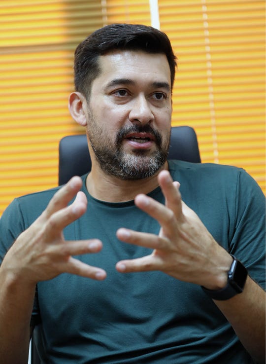 Um homem de cabelos escuros e barba curta grisalha aparece em primeiro plano, gesticulando com as mãos enquanto fala. Ele veste uma camiseta verde-escura e usa um relógio inteligente no pulso esquerdo. O homem está sentado em uma cadeira de escritório preta, e ao fundo há uma persiana de cor alaranjada que filtra a claridade, criando um ambiente de conversa ou entrevista.