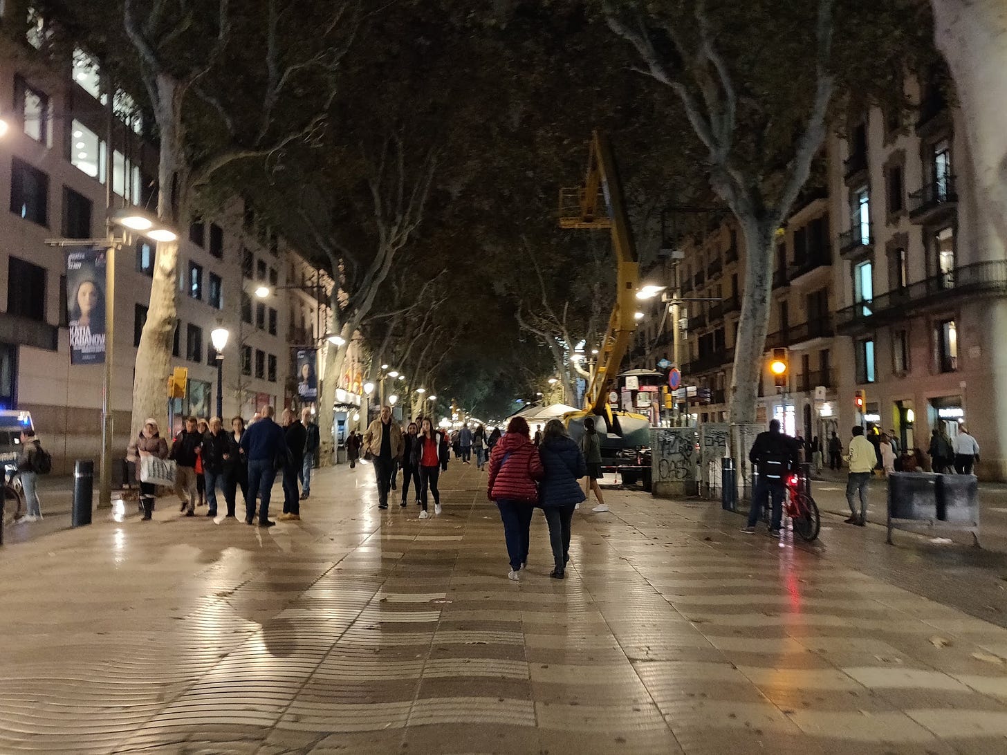 La Rambla in Barcelona at night, crowds of people walking under tall plane trees lit by street lamps.