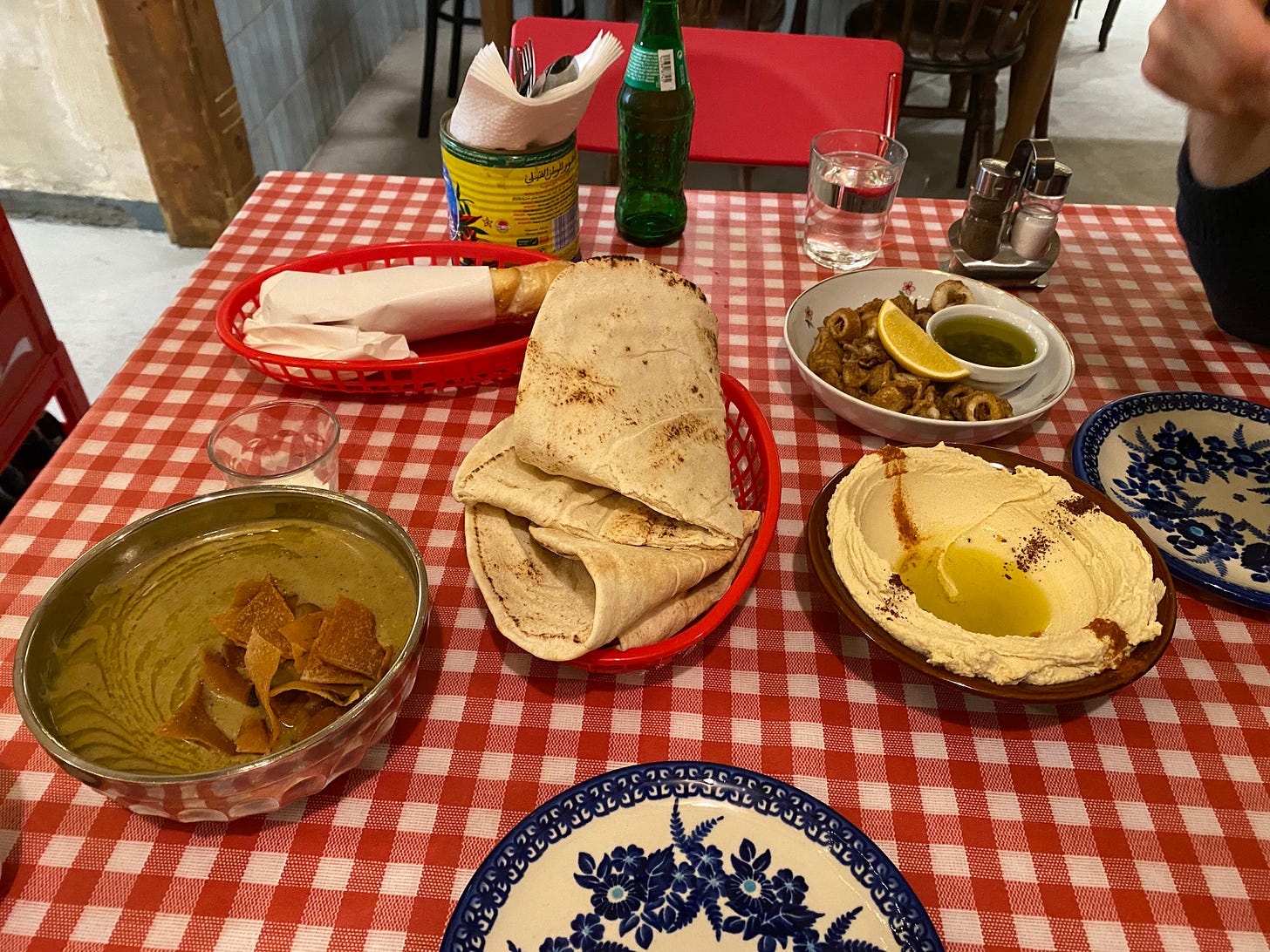 Many plates and bowls of food on a simple checkered table. There are breads, dips and olives. It looks homely.