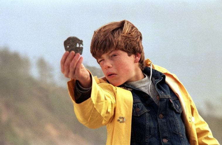 Gif dal film "I Goonies", con un bambino con giubbino di jeans e giubbetto impermeabile giallo, mentre chiude un occhio per guardare dentro una specie di moneta a forma di teschio.