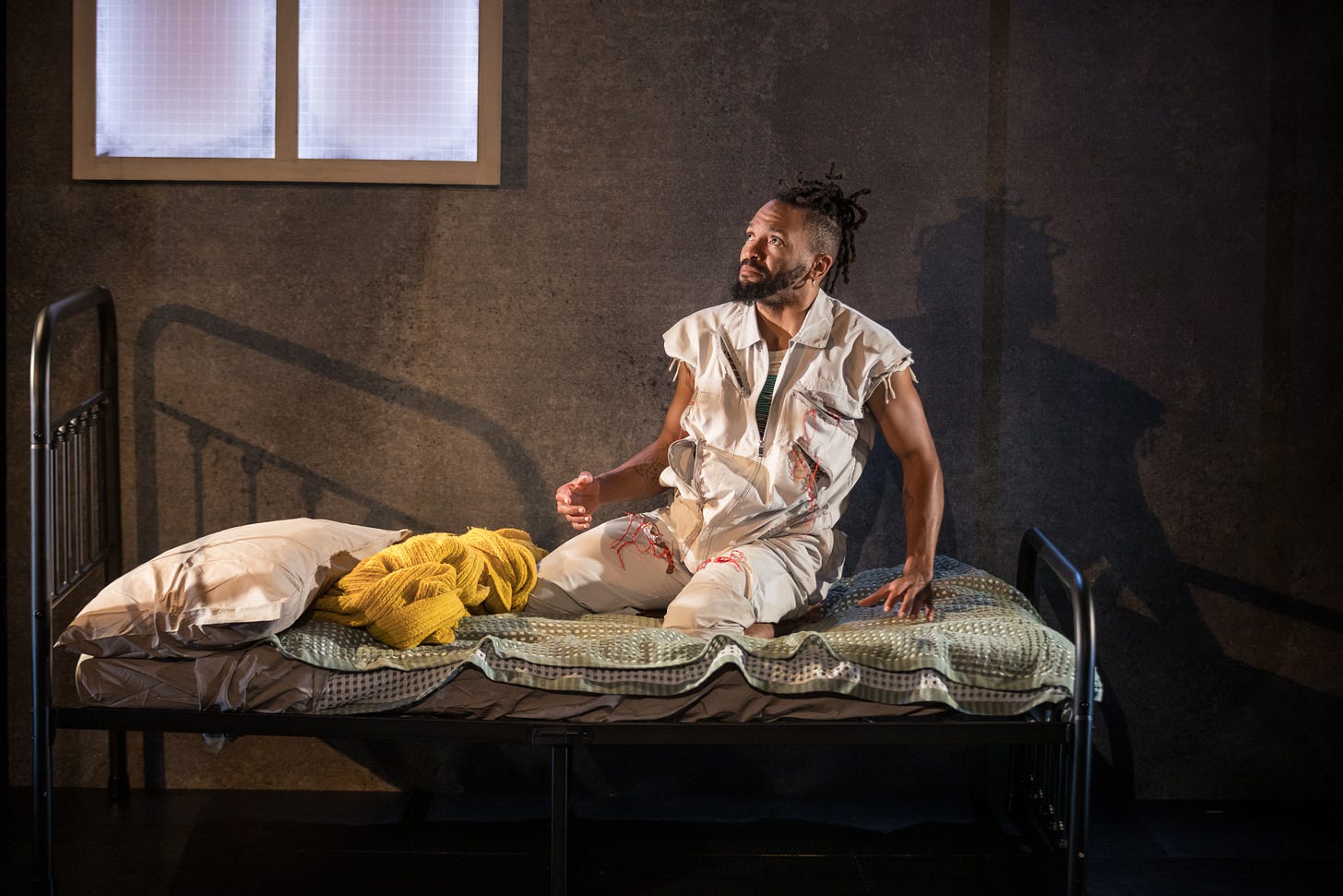 A black man with hair in an up-do and a beard kneels on a bed, wearing a white jumpsuit.
