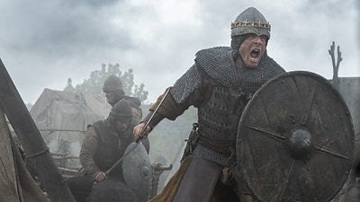 James Norton in character as King Harold wearing body armour and charging at speed on horseback. He is holding a shield and sword.