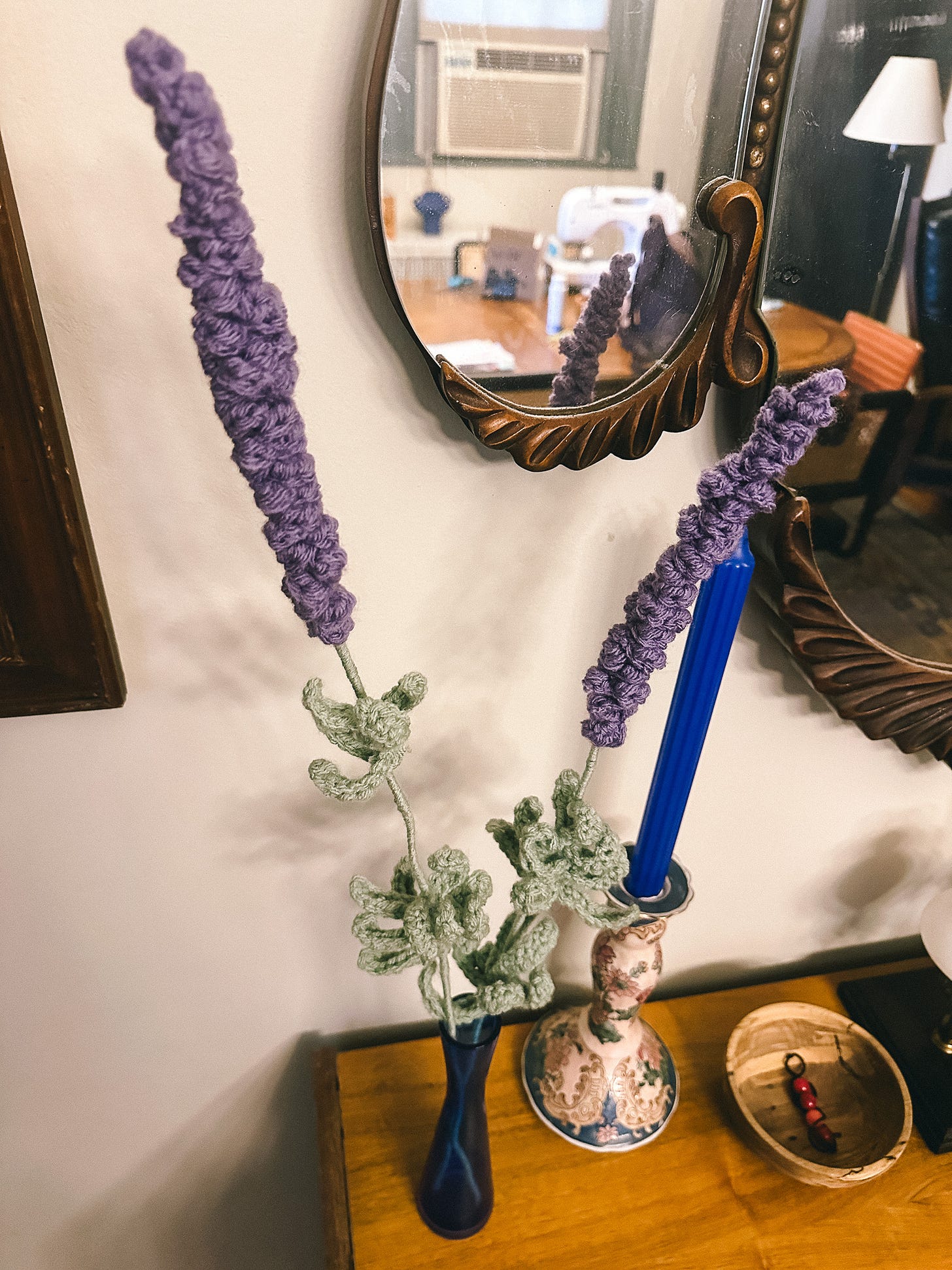 two lavender stalks made of yarn in a vase on a table next to a blue candle and a wooden bowl, with a large mirror behind them reflecting a dining room table that has a sewing machine on it