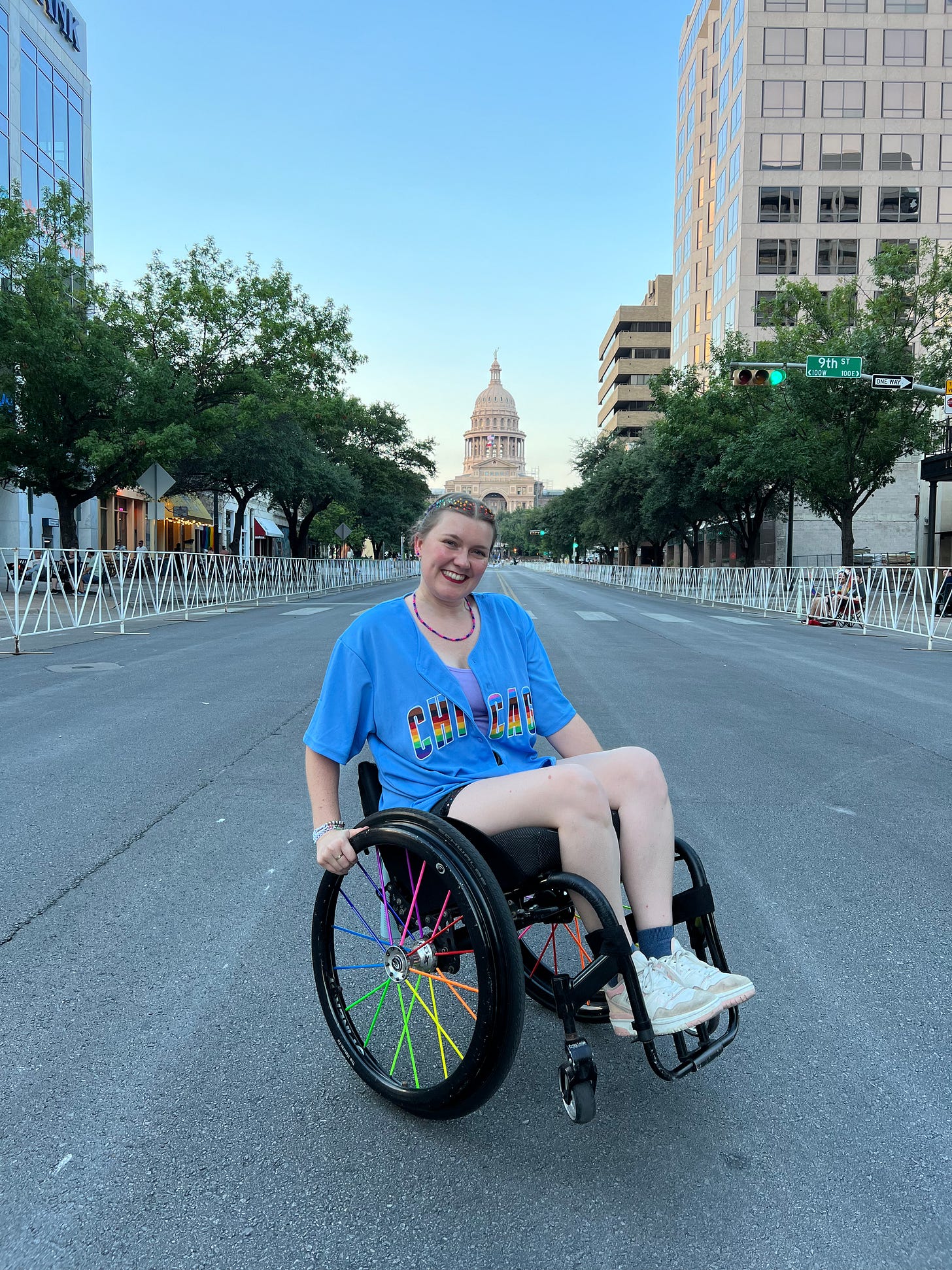A person in a wheelchair smiles brightly on an empty street leading to the Texas State Capitol. They wear a blue t-shirt with "Chicago" in rainbow colors, matching their wheelchair's colorful spokes and rainbow-bejeweled hair. The scene is framed by tall buildings, trees, and a clear blue sky.