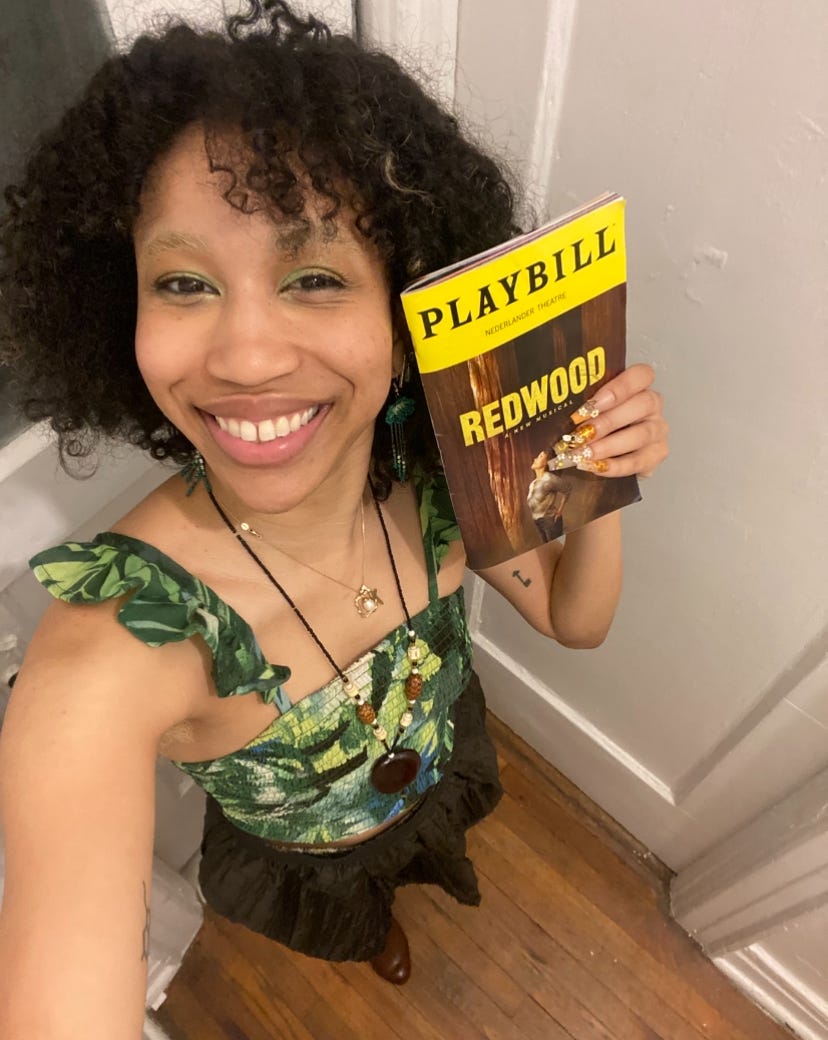 Kiana, a light skin woman with curly dark hair is wearing a leaf patterned shirt and a brown skirt to match the redwood playbill 