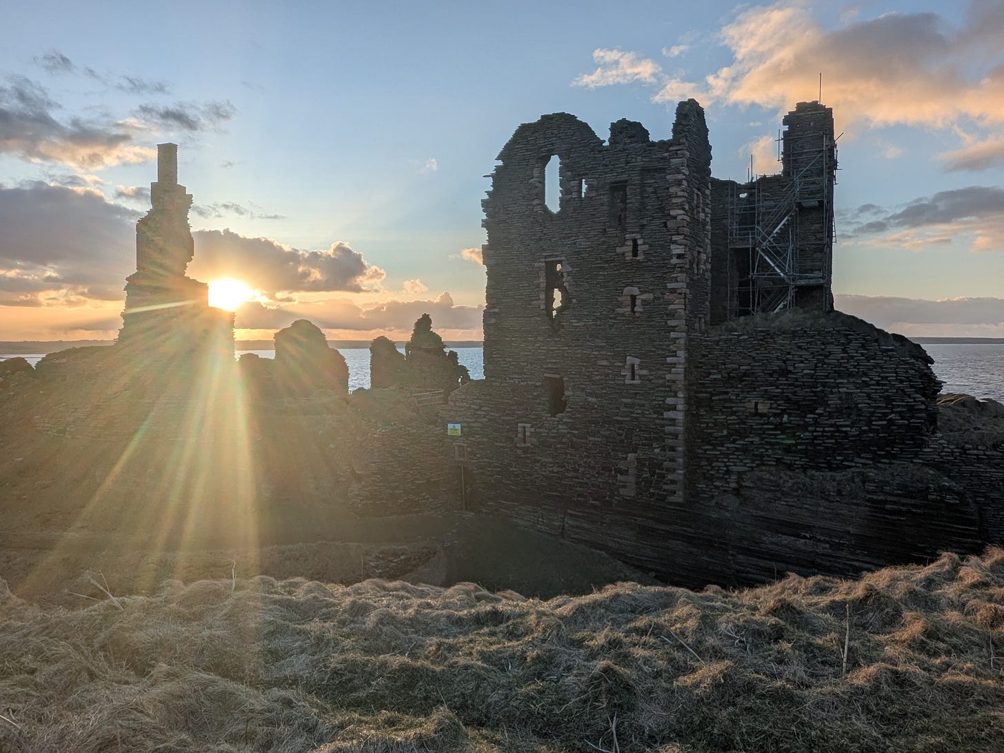 A landscape photograph of a ruined stone castle on a grassy clifftop at sunset. The sun is shining brightly through a gap in the ruins on the left, creating a starburst effect. The ocean is visible in the background under a partly cloudy, golden sky.