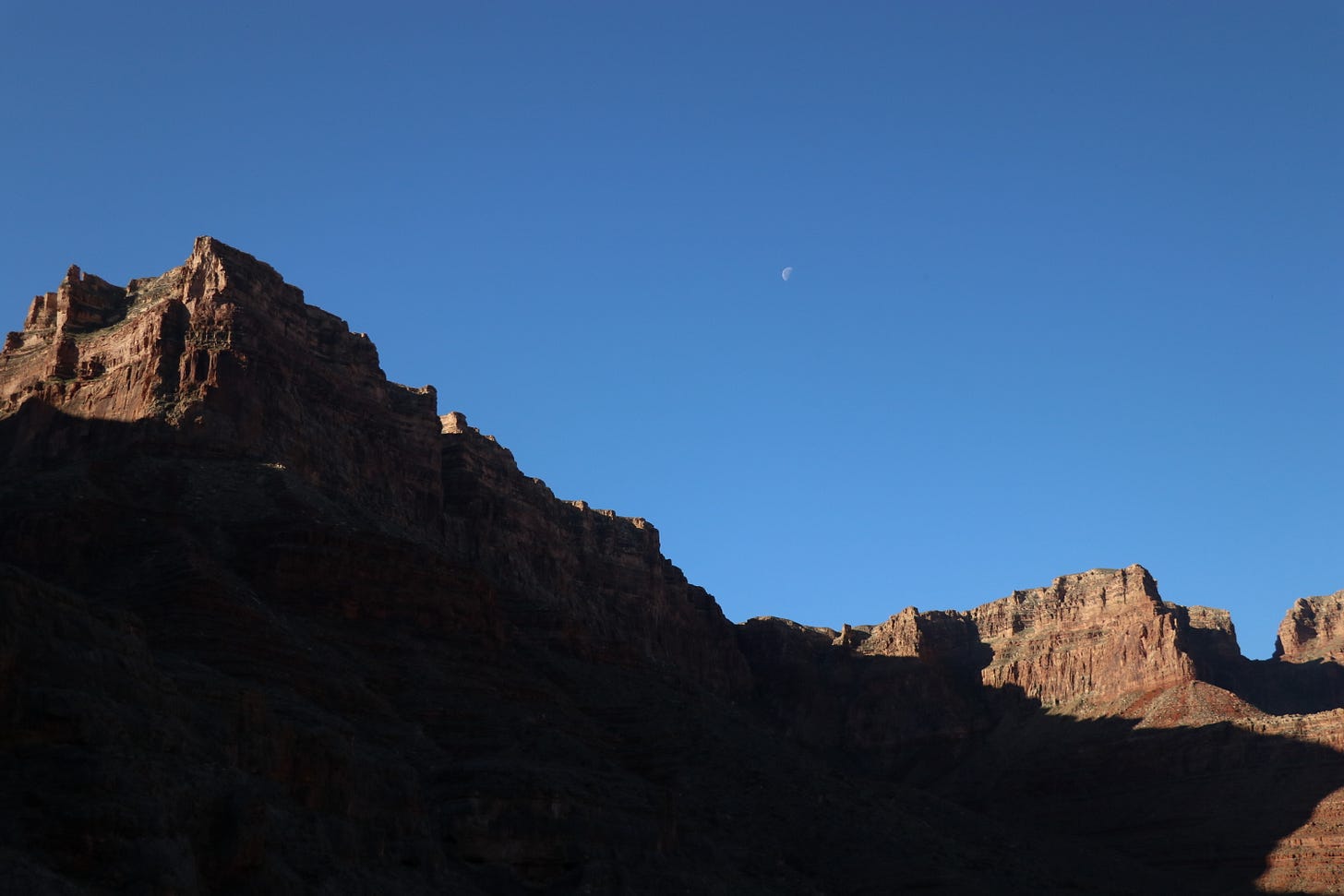 A half moon sits in a dimming evening sky above the rim of the Grand Canyon, half in shadow and half in light