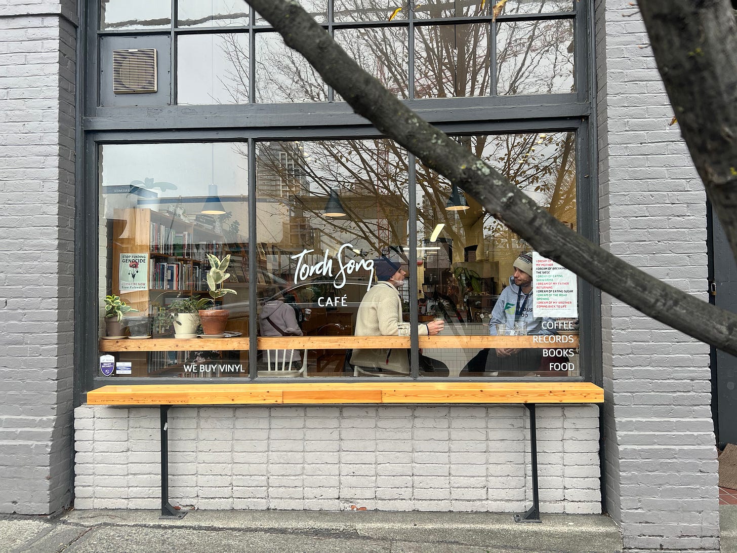 The exterior of a cafe, with a big window. Inside the window you can see two mean eating and talking, and several bookshelves.