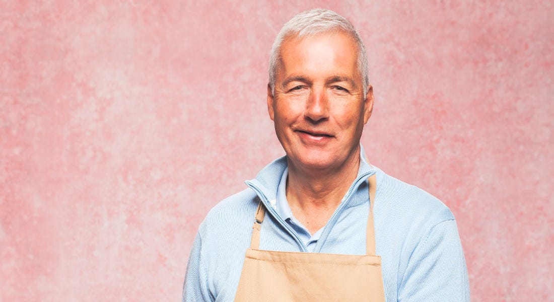 A man wearing a blue shirt and brown apron, in front of a pink background A man wearing a blue shirt and brown apron, in front of a pink background