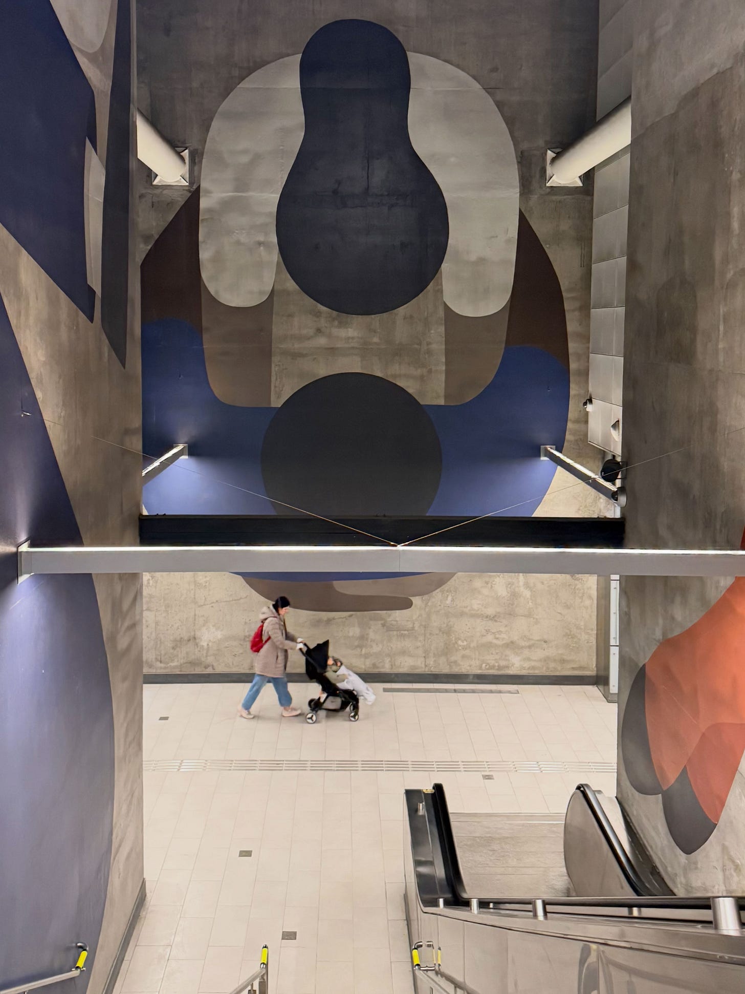 A woman pushing a stroller walks at the bottom of a long flight of stairs. They are drarfed by a large piece of public art