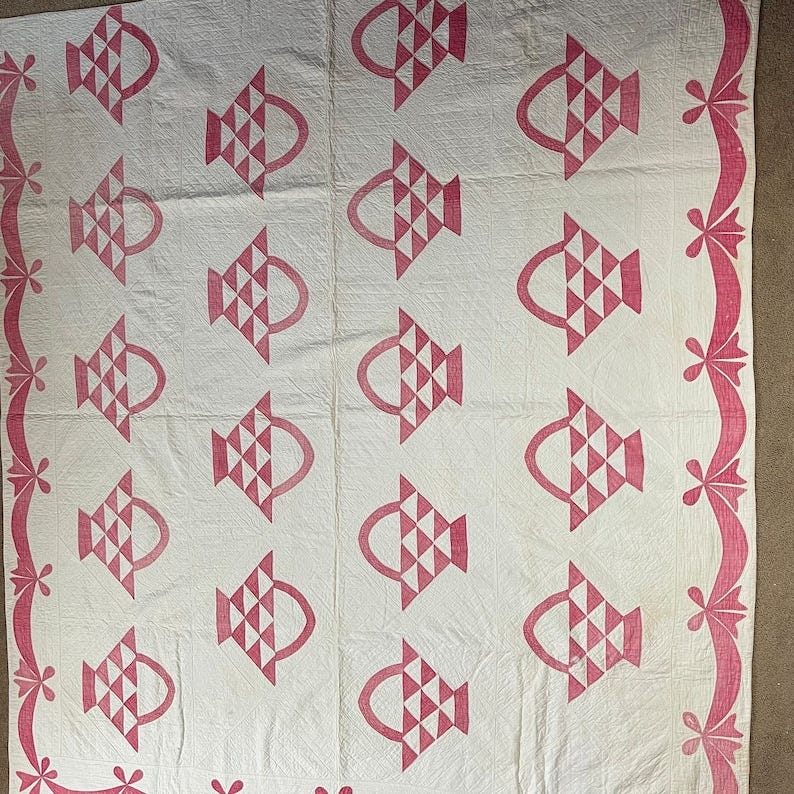 May include: A white quilt with a pink basket pattern. The quilt has a pink border with a floral design. May include: A white quilt with a pink basket pattern. The quilt has a pink border with a floral design.