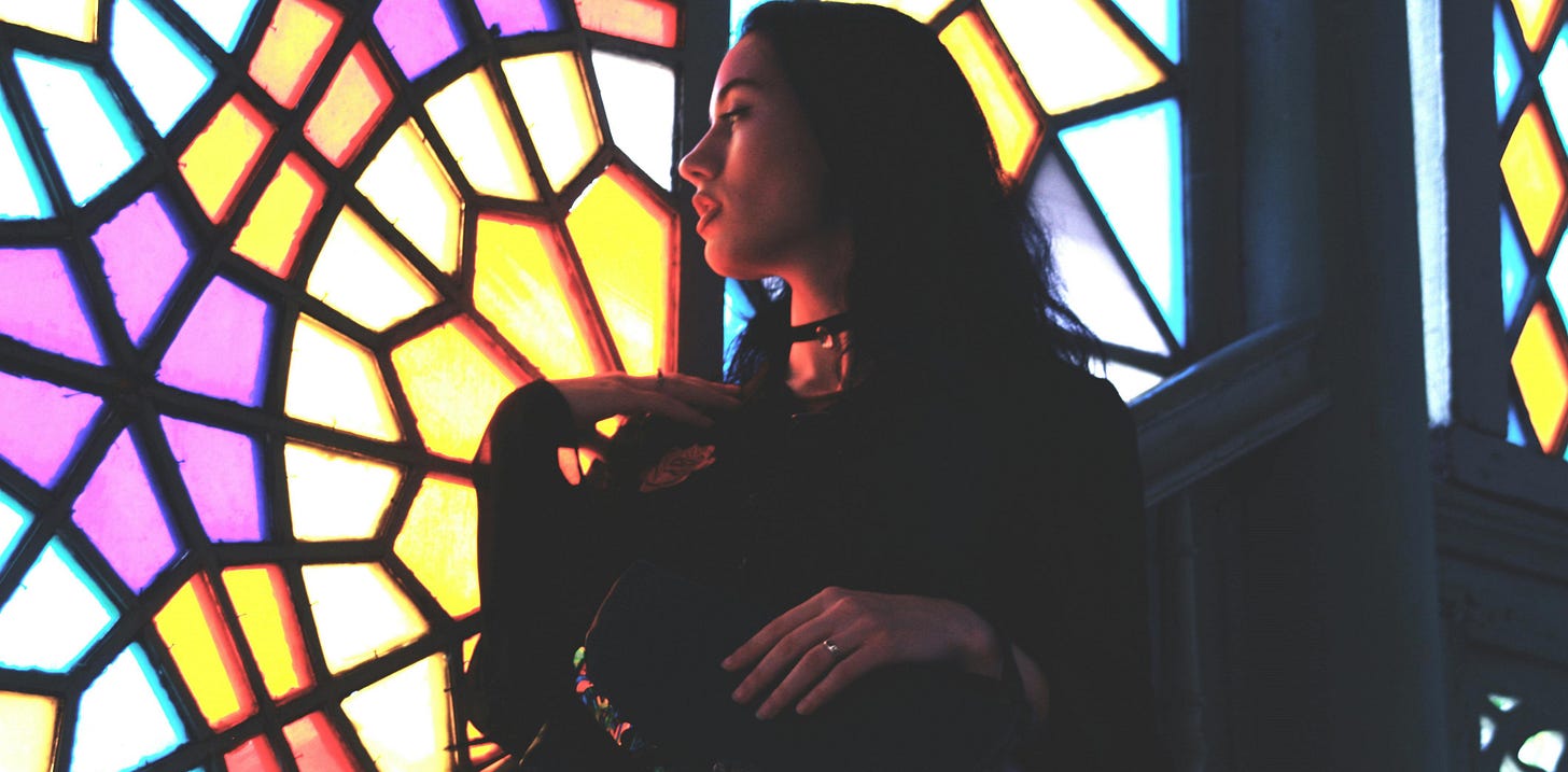 A woman looks out through a yellow, purple, and blue stained glass window.