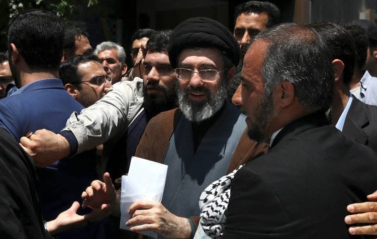 A group of men gather in conversation outdoors, with a cleric in a black turban and glasses at the center holding papers. (Photo Credit: AP Photo/ Vahid Salemi)