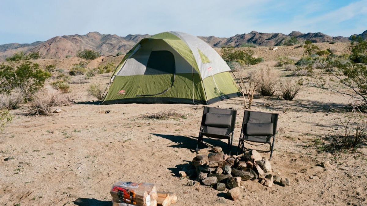 Image: Two folding chairs propped infront of an unlit campfire ring with a green tent behind and the brown sandy desert and low mountains in the background.