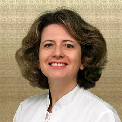 Uma mulher aparece em um retrato de fundo neutro bege, olhando diretamente para a câmera e sorrindo. Ela tem cabelos curtos, castanhos com mechas mais claras, levemente ondulados e na altura do queixo. Veste um jaleco branco, sugerindo atuação na área da saúde. A imagem é bem iluminada e enquadrada do peito para cima, transmitindo uma aparência profissional e acolhedora. Uma mulher aparece em um retrato de fundo neutro bege, olhando diretamente para a câmera e sorrindo. Ela tem cabelos curtos, castanhos com mechas mais claras, levemente ondulados e na altura do queixo. Veste um jaleco branco, sugerindo atuação na área da saúde. A imagem é bem iluminada e enquadrada do peito para cima, transmitindo uma aparência profissional e acolhedora.