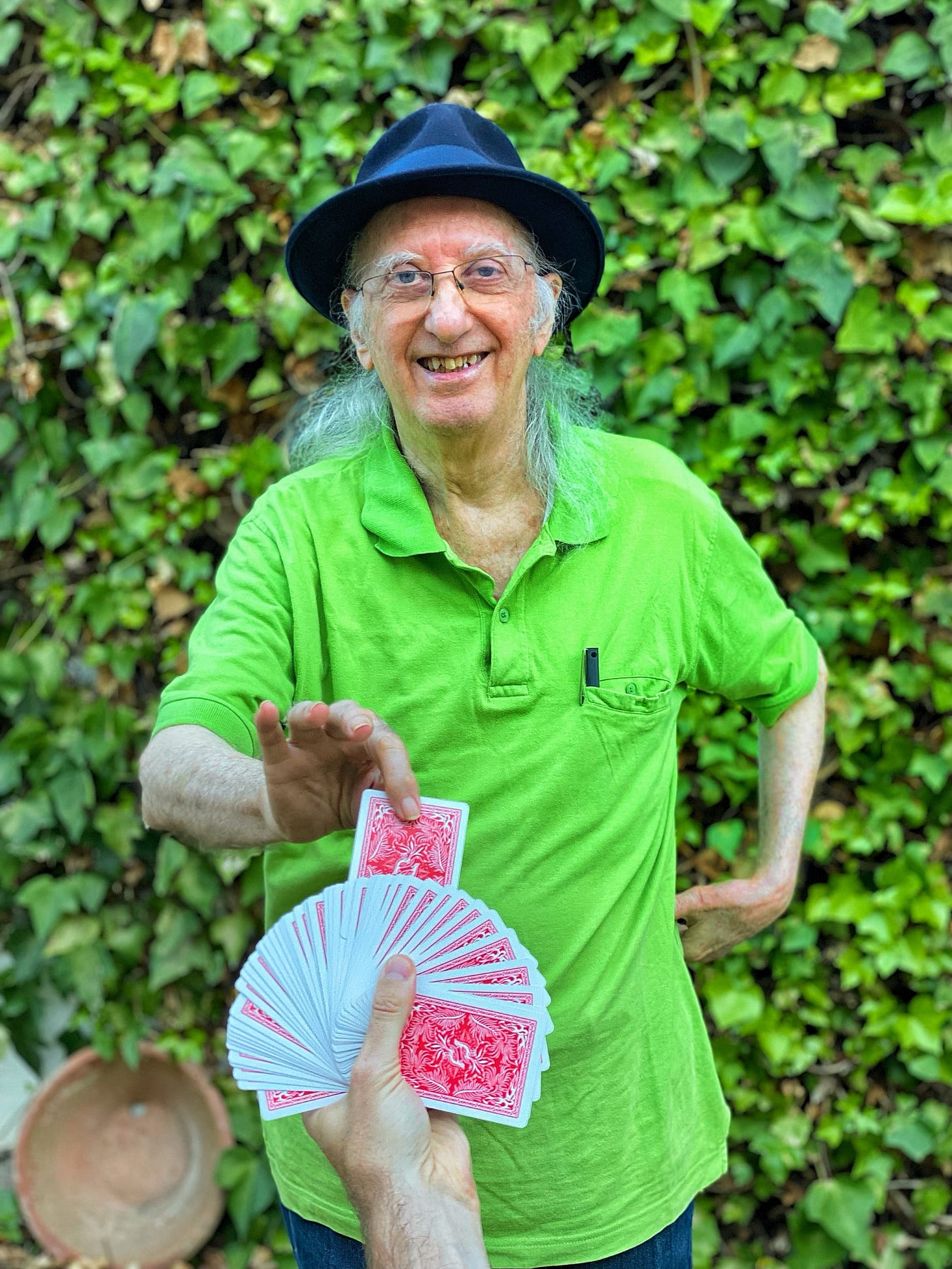 Magician Juan Tamariz, wearing a bright green polo shirt and a dark hat, smiles widely as he reaches out to select a card from a fanned deck of red playing cards held toward him. He is standing outdoors in front of a lush wall of green ivy. Magician Juan Tamariz, wearing a bright green polo shirt and a dark hat, smiles widely as he reaches out to select a card from a fanned deck of red playing cards held toward him. He is standing outdoors in front of a lush wall of green ivy.