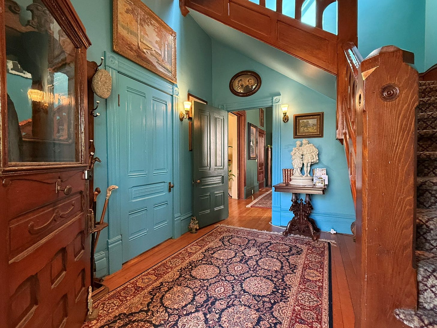 A view of the foyer taken from the threshold of the front door. A hall tree at left collects canes and hats. There are two doors: one into the dining room (open) and another into the parlor (closed). There is a shairway at right.