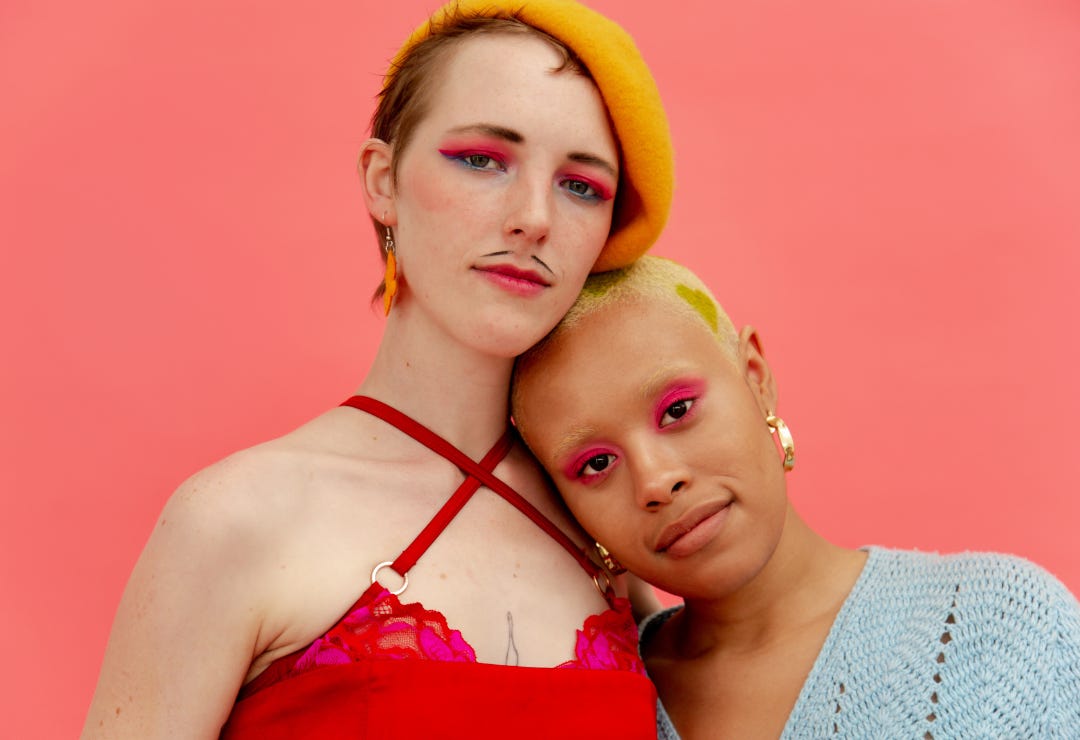 black woman with shaven yellow hair and red eyeshadow leans on the shoulder of a white man wearing red eyeshadow and a red dress black woman with shaven yellow hair and red eyeshadow leans on the shoulder of a white man wearing red eyeshadow and a red dress