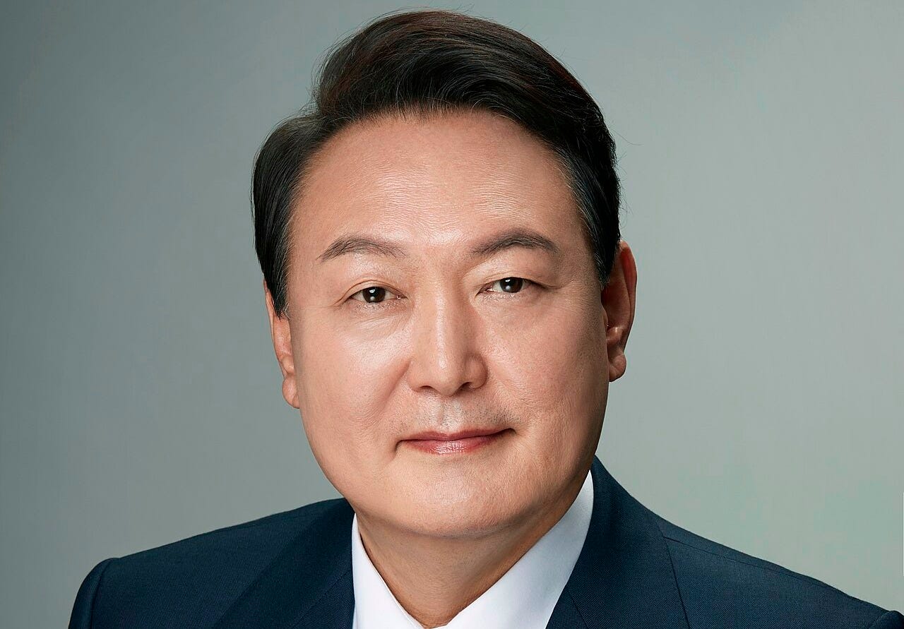 Head shot of Yoon in front of a gray background, wearing a dark suit jacket with a white shirt and a blue and pink patterned necktie.