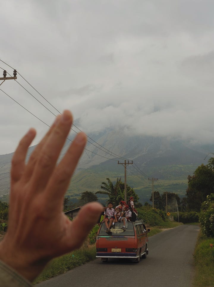 A van crosses a strech of the jungle through the fog. A hand says goodbye to a schoolbus with kids siting on the roof of their bus. In the background is the active volcano Mount Sinabung