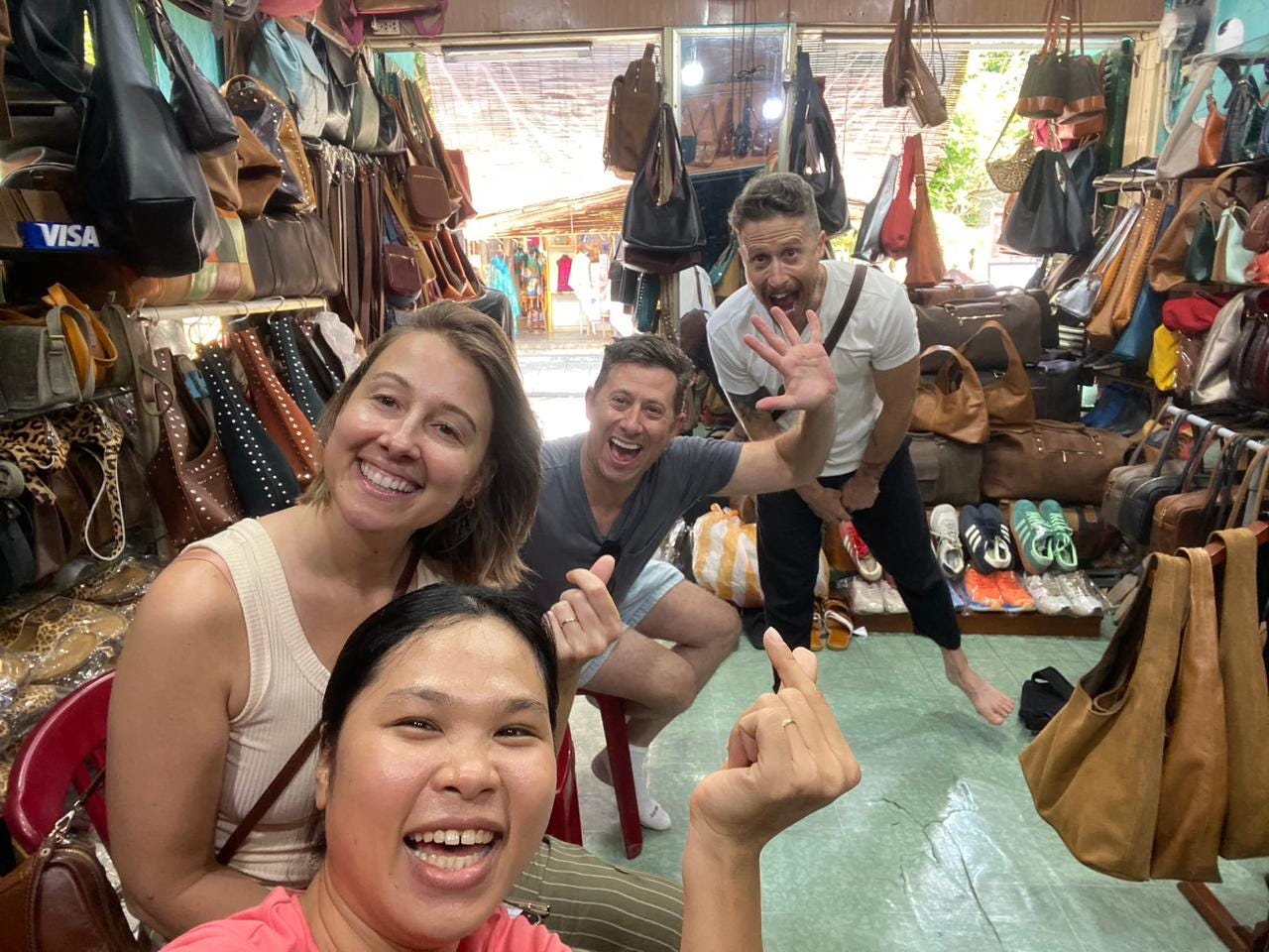 Four friends grinning and waving inside a small Vietnamese leather shop, surrounded by rows of handmade bags, giving a mix of peace signs and playful gestures