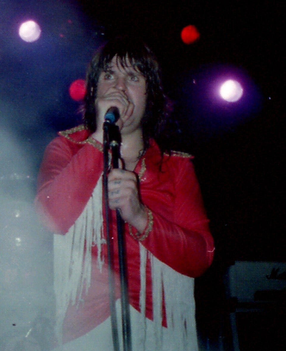 Ozzy on stage, singing into a microphone. He’s wearing a red shirt with white fringes.