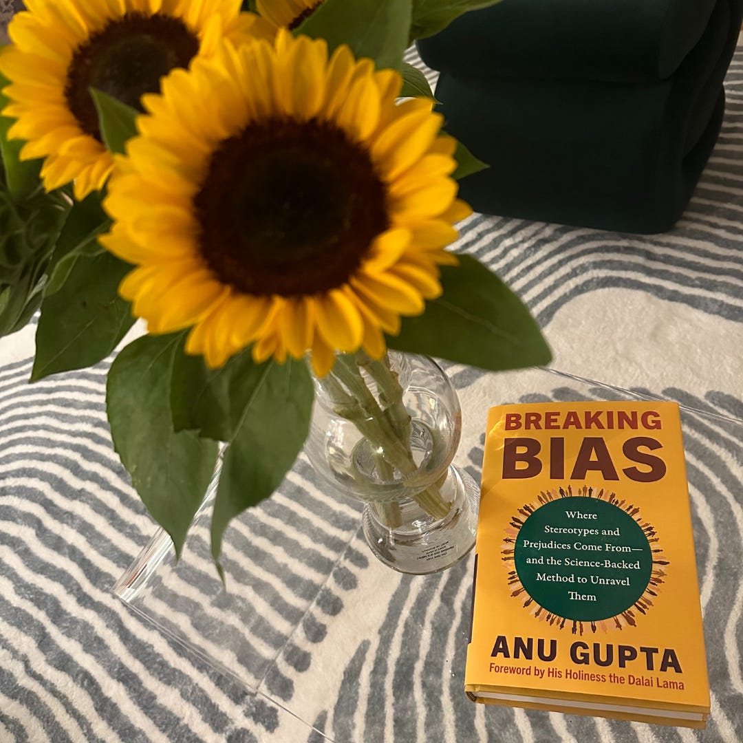 A yellow book on a glass table next to a vase of sunflowers with meditation cushions nearby.