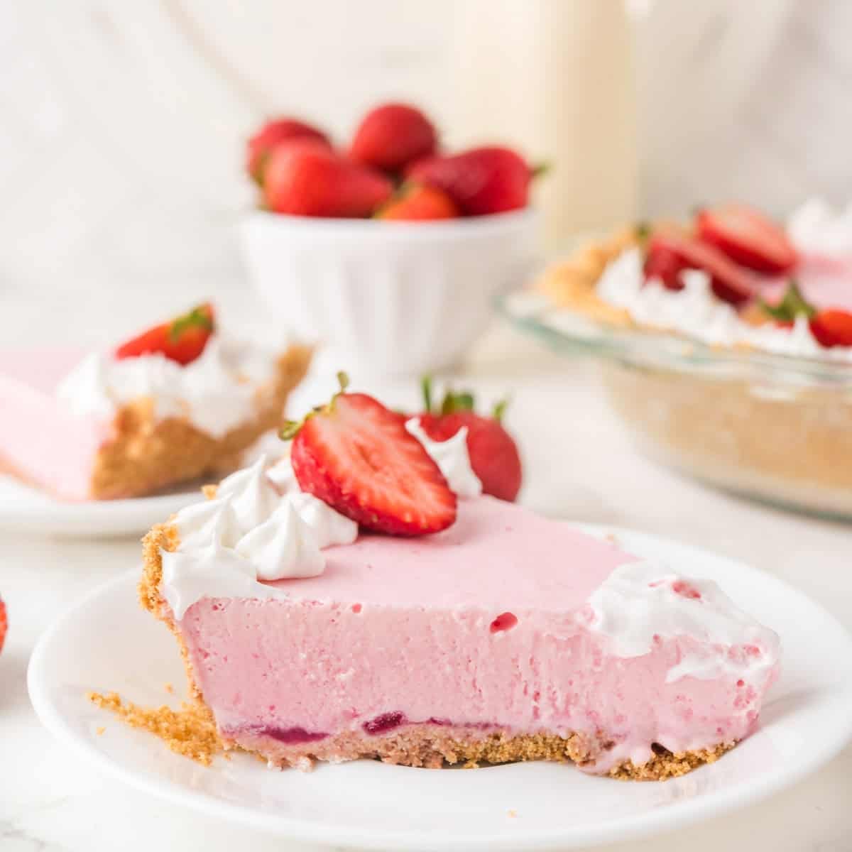 A slice of pink strawberry cream pie with a graham cracker crust, whipped cream, and fresh strawberry slices on top sits on a white plate. Whole strawberries and more pie are in the background.
