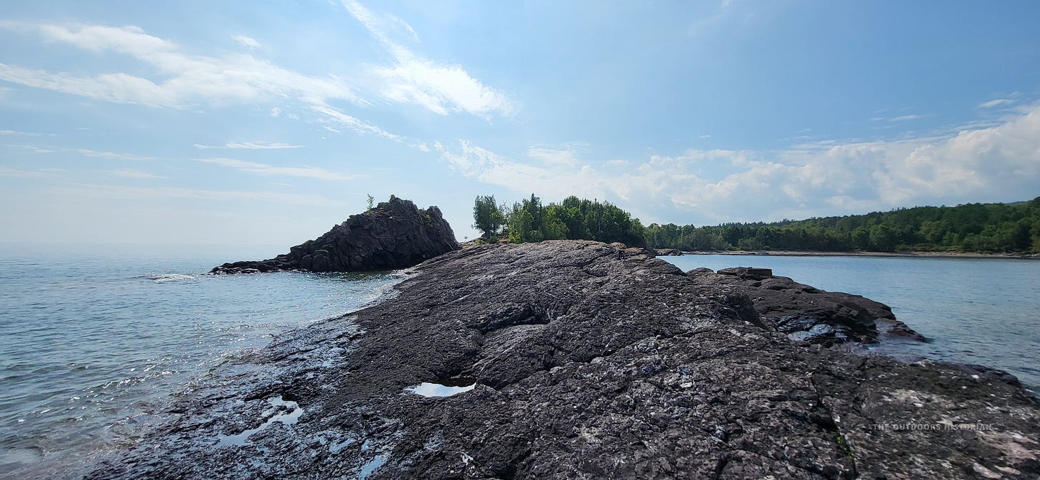 A close-up of dark, jagged volcanic rock (basalt) on the shore of Lake Superior. The rock layers are visibly tilted toward the water, showing ancient geological movement.