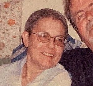 A white woman looks off past the camera, smiling, on a couch with a man.
