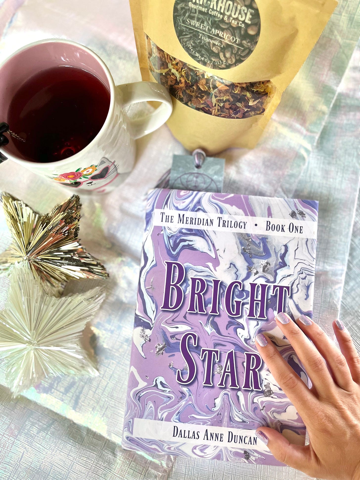 Indie fantasy author Dallas Anne Duncan's hand grazes the marbled cover of "Bright Star", her first book. The book lays on top of an iridescent tablecloth and is surrounded by tinsel stars, a hot tea mug, and a bag of artisan tea. Indie fantasy author Dallas Anne Duncan's hand grazes the marbled cover of "Bright Star", her first book. The book lays on top of an iridescent tablecloth and is surrounded by tinsel stars, a hot tea mug, and a bag of artisan tea.