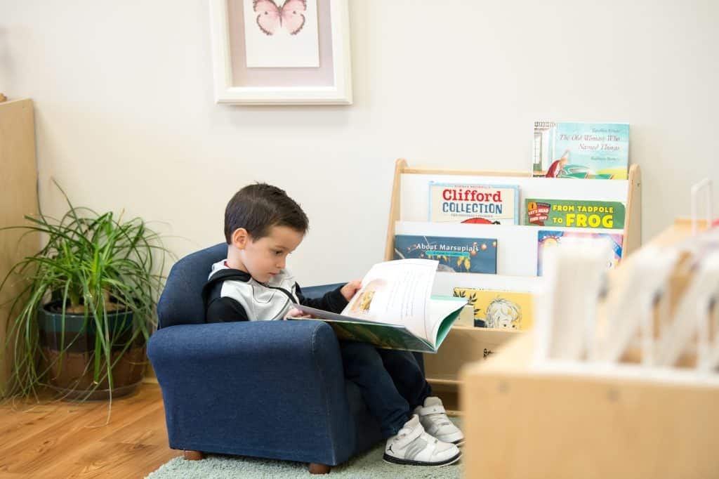Child engaged in reading a book on a small blue armchair in a home learning space.
