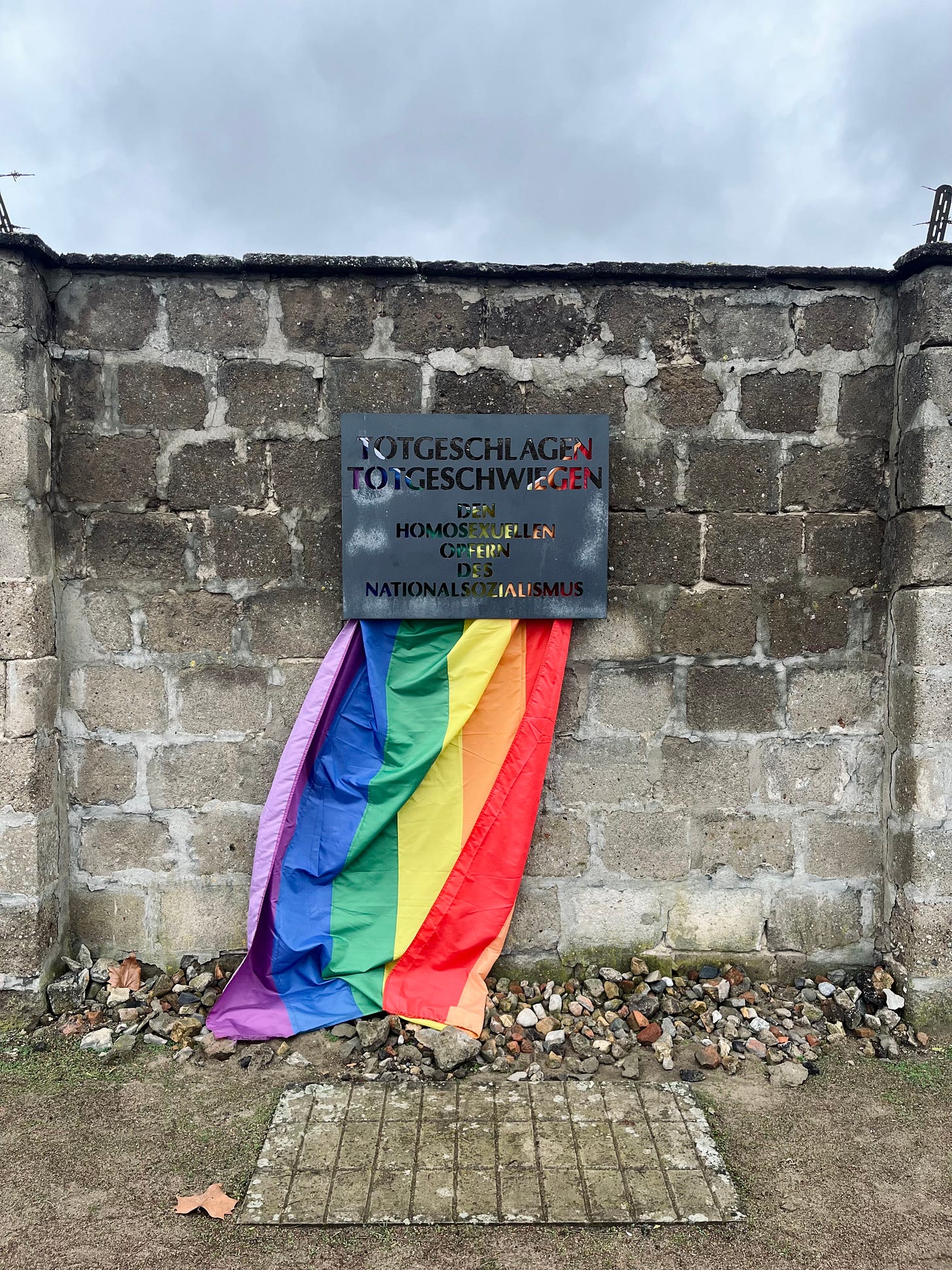 A rainbow flag hangs from a metal sign on a brick wall. the sign reads "totgeschlagen totgeschwiegen" 