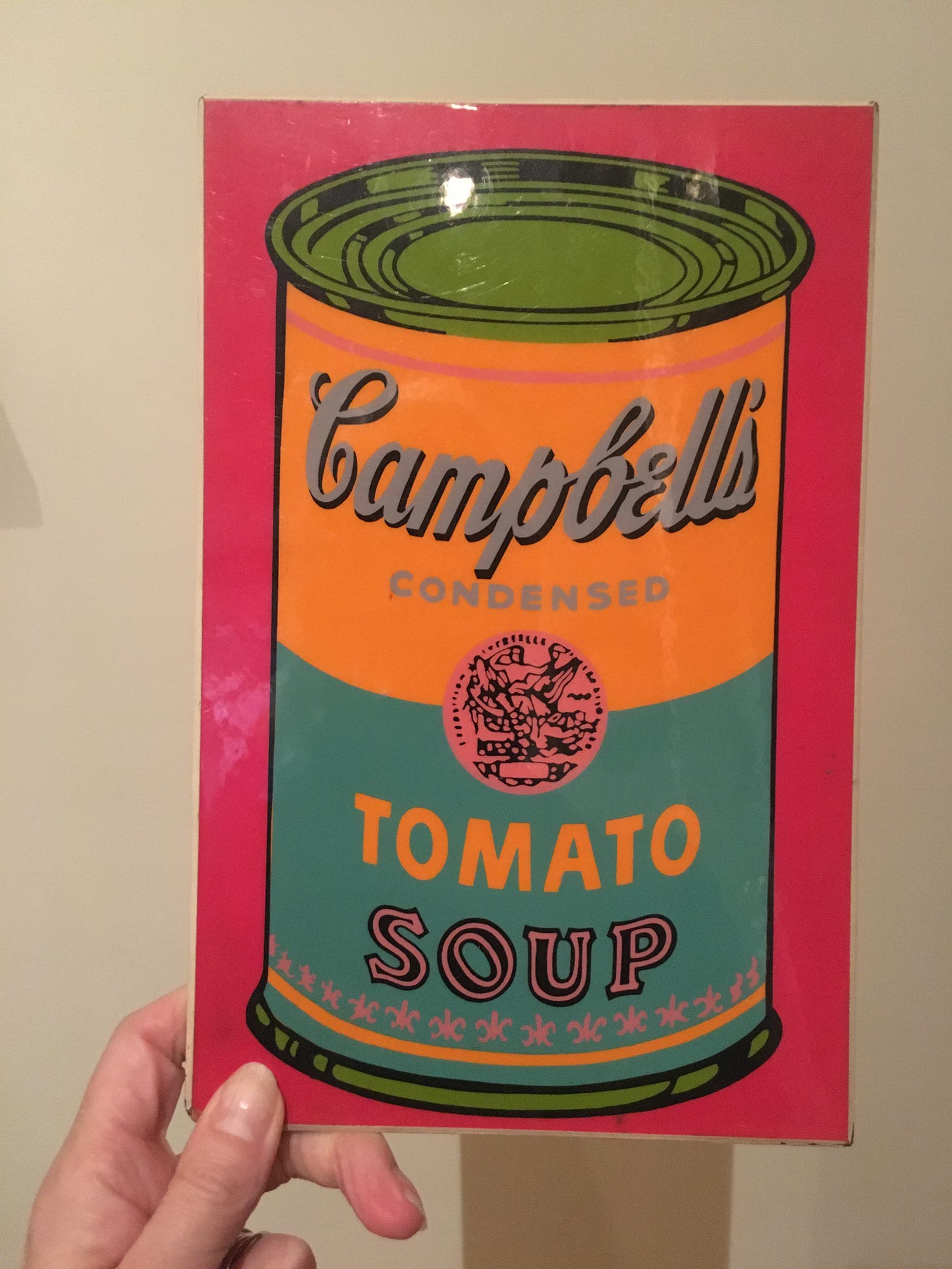 pink and orange reproduction of a tomato Campbell's soup hand held by a visible hand with thumb on front.