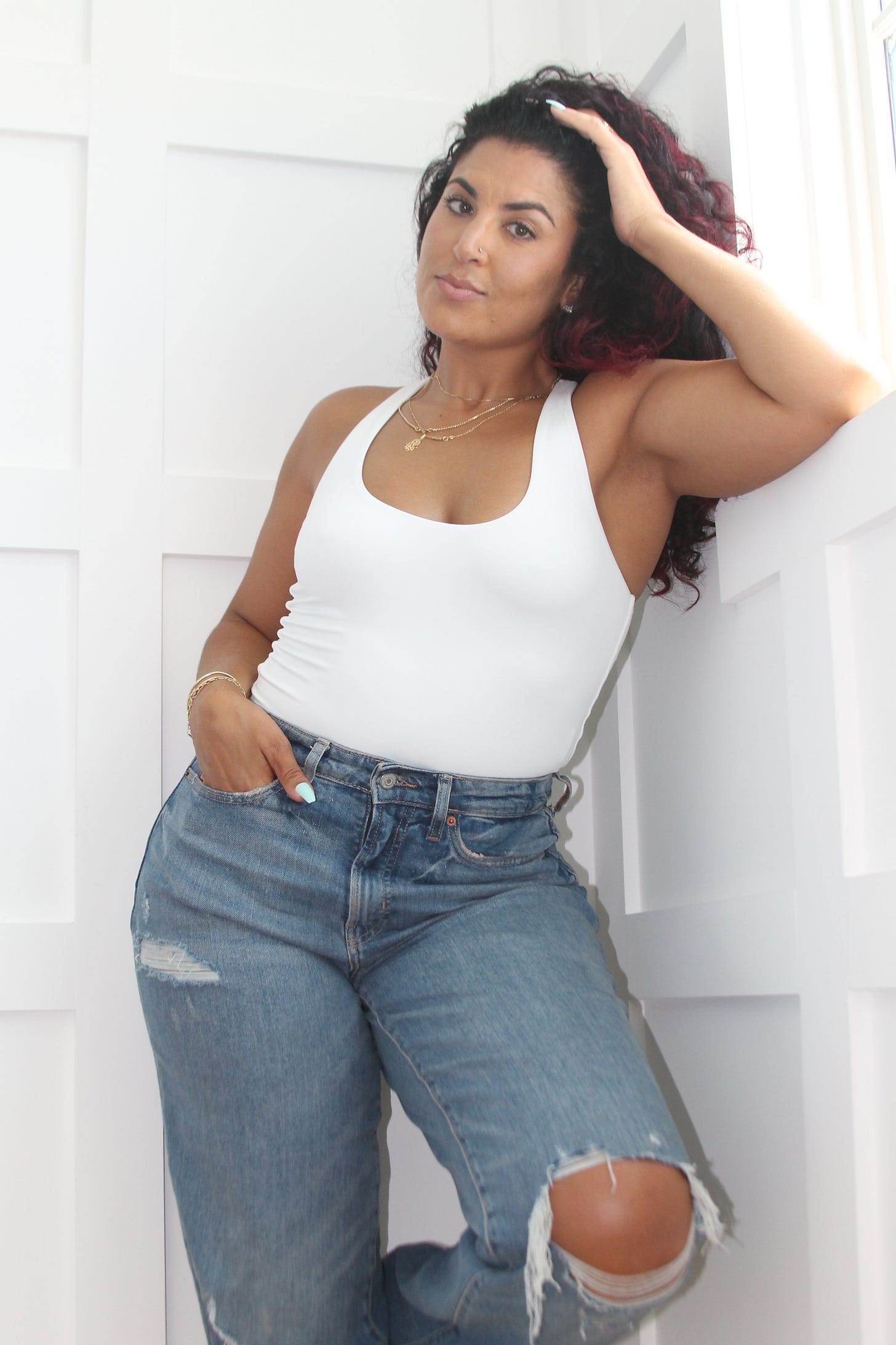 Woman standing in a white body suit and blue jeans leaning against a white wall with one hand in her pocket and the other resting behind her head.