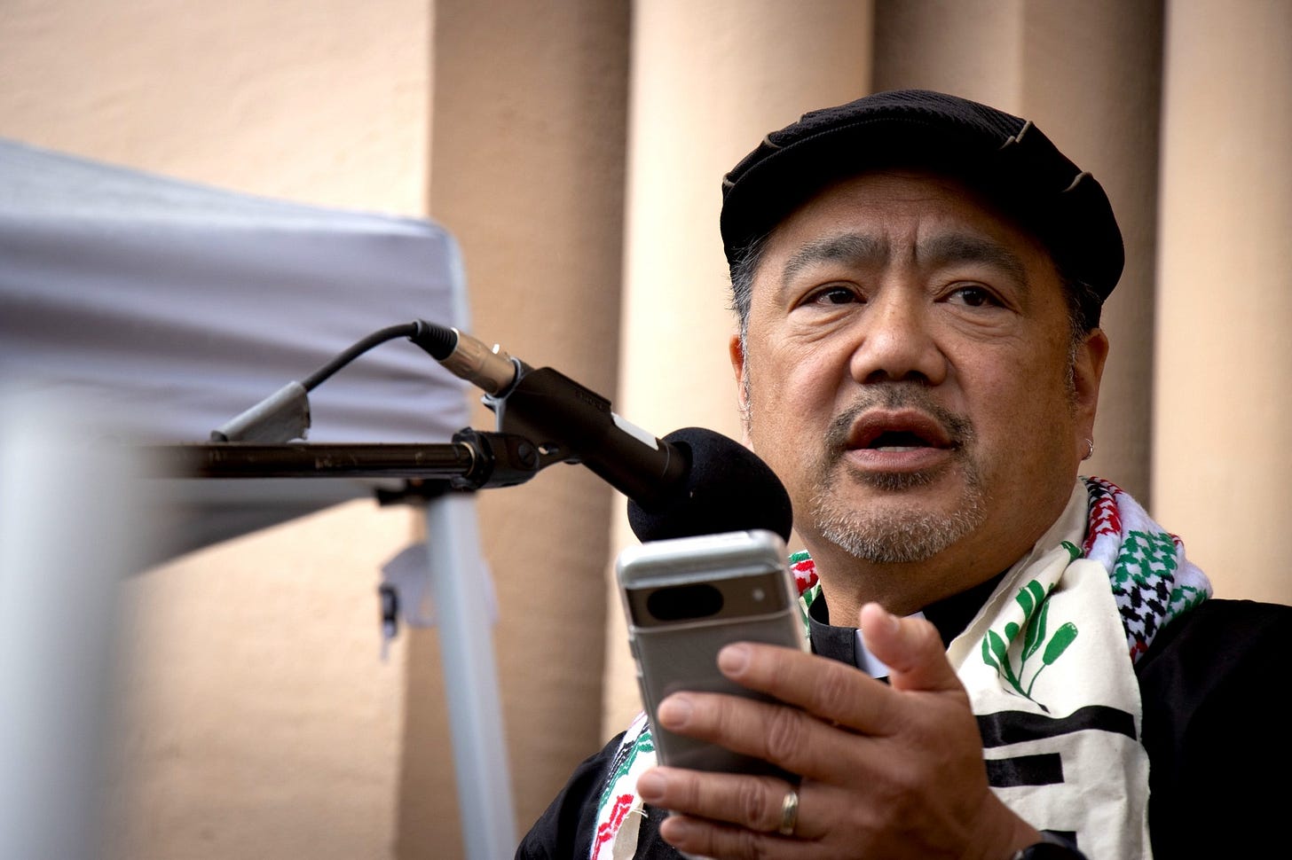 Bruce is at a microphone reading remarks from his phone. He is wearing a black cap, and a red, green, and black kefiah around his shoulders
