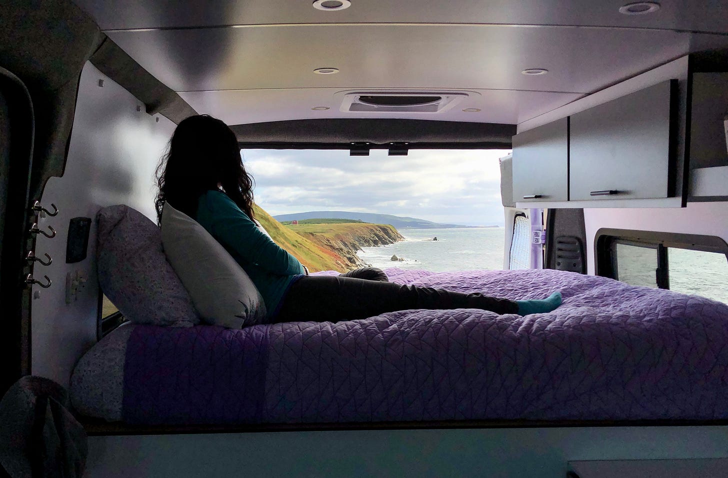 A woman on a bed in a campervan looking at the sea through open back doors. A woman on a bed in a campervan looking at the sea through open back doors.