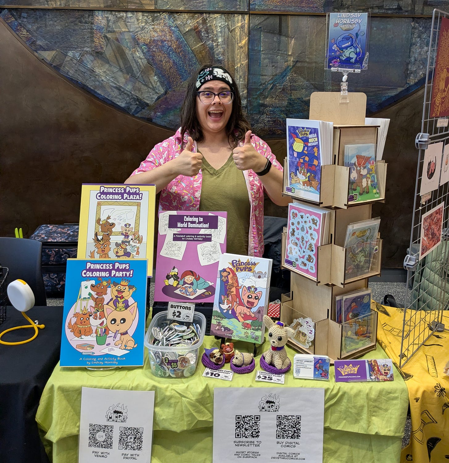 Lindsay Hornsby, a dark haired cartoonist with glasses, gives double thumbs up behind a table at Alaska Mini-Con 2026. She's selling lots of comics, minis, stickers and more wit her comics and art on them!