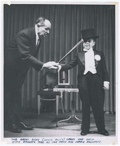 An older magician onstage next to a young boy magician in top hat and tails. An older magician onstage next to a young boy magician in top hat and tails.
