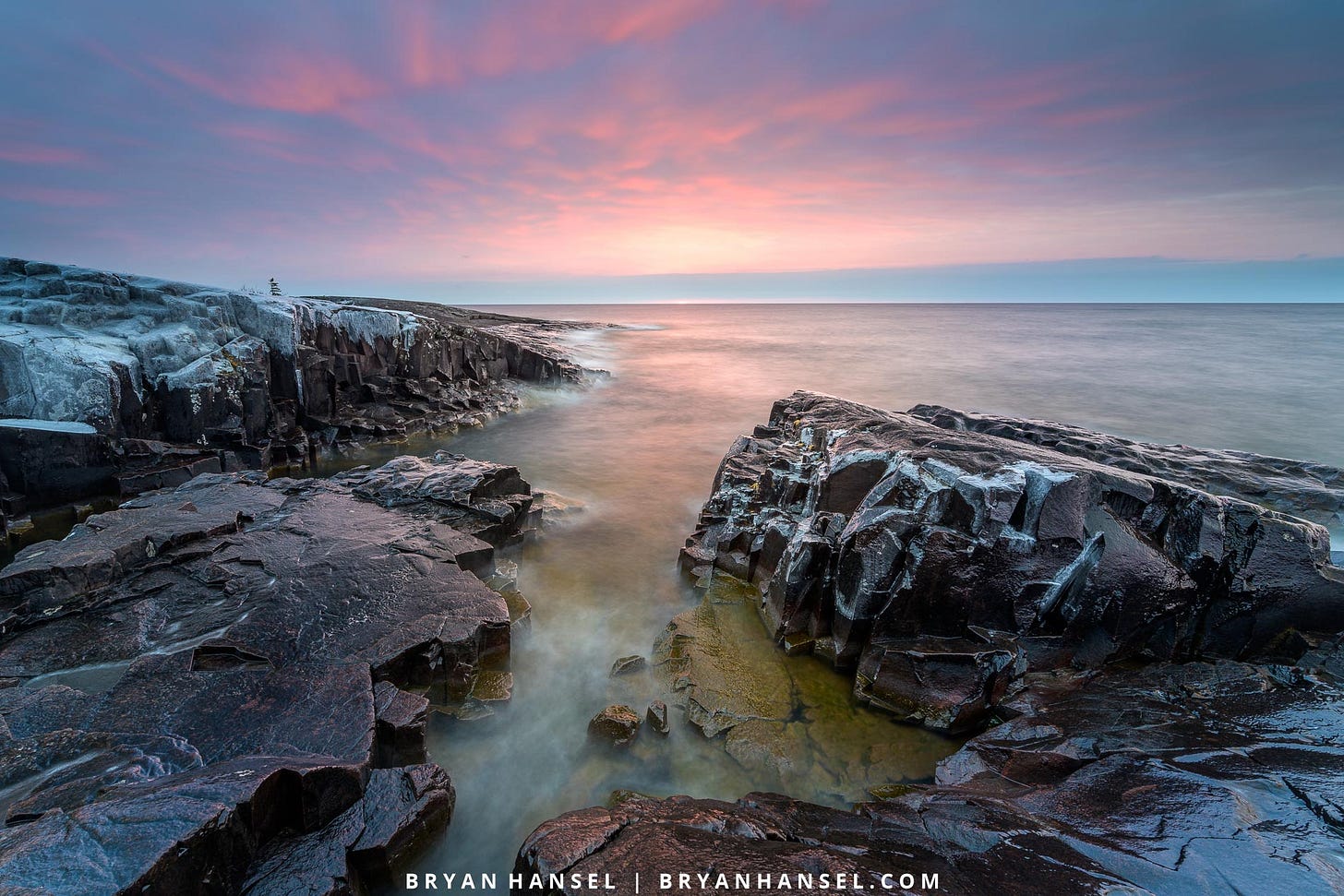 A photo of sunrise over Lake Superior. A basalt shoreline has a crack/slot cut right through it and it aims at a purple and pink sunrise.